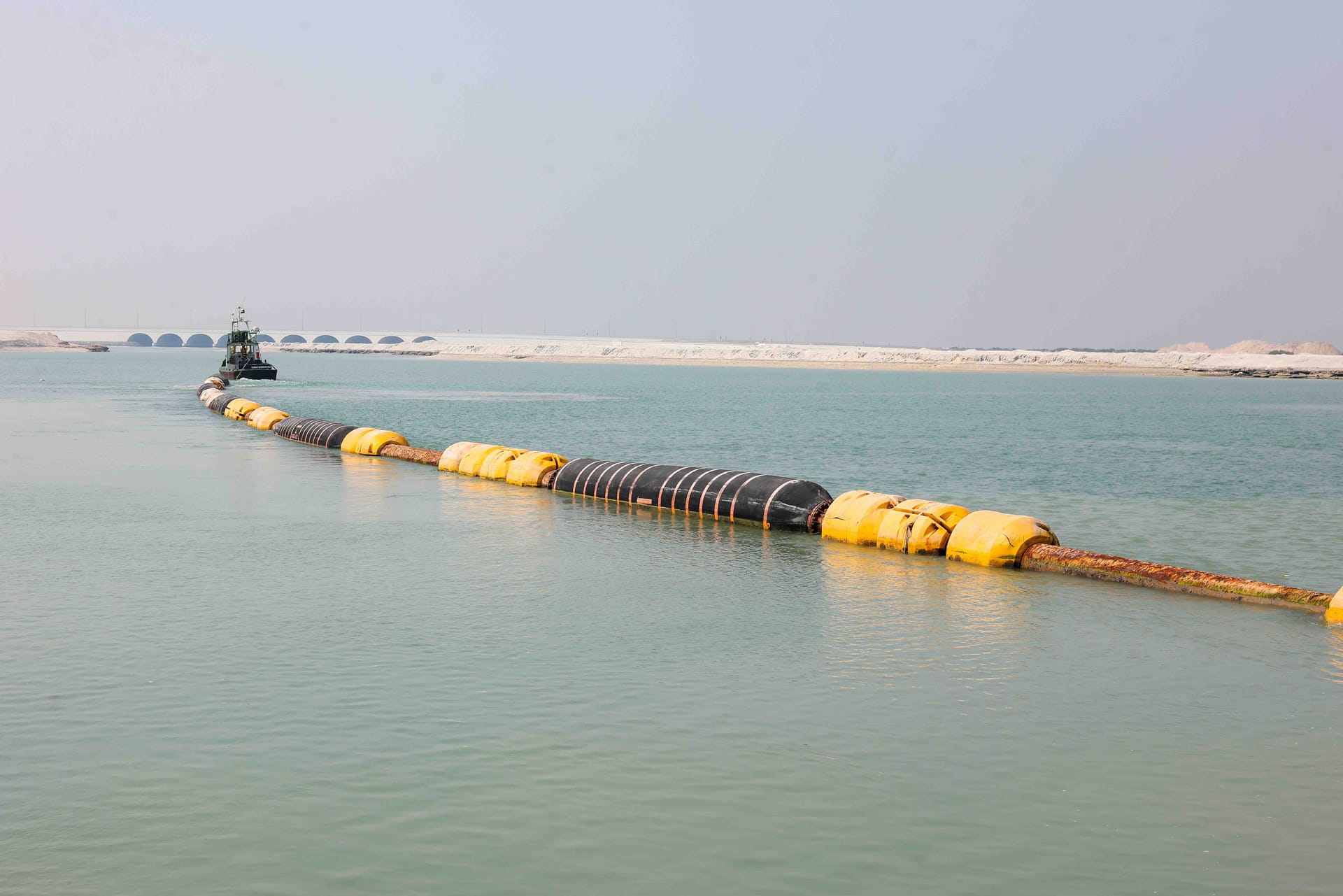 Floating dredge discharge pipeline on yellow floats stretching across a channel to a distant workboat.