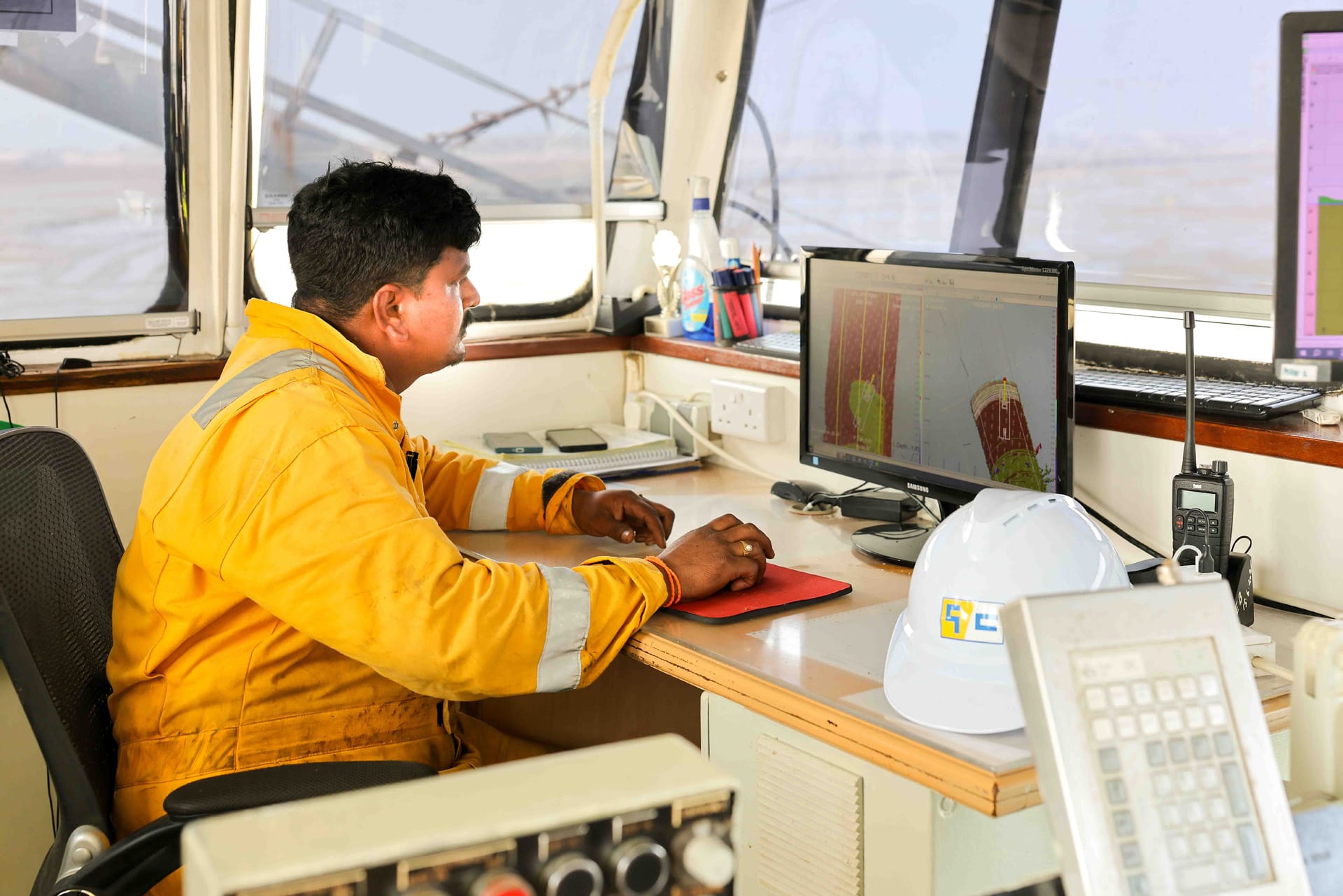Surveyor in a yellow coverall reviewing survey software onboard, with hard hat and handheld radio nearby.